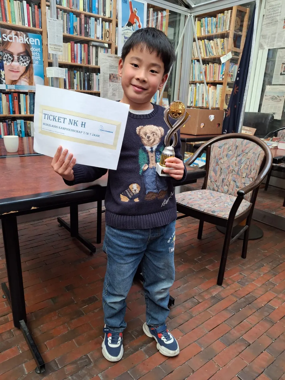 Een jonge schaker toont trots zijn ticket en een beker tussen de boekenkasten